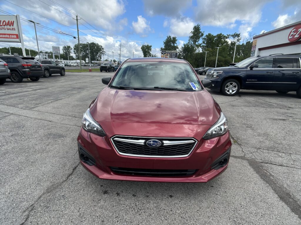 Front view of a red Subaru SUV in a parking lot with other cars and a dealership building in the background under a partly cloudy sky.