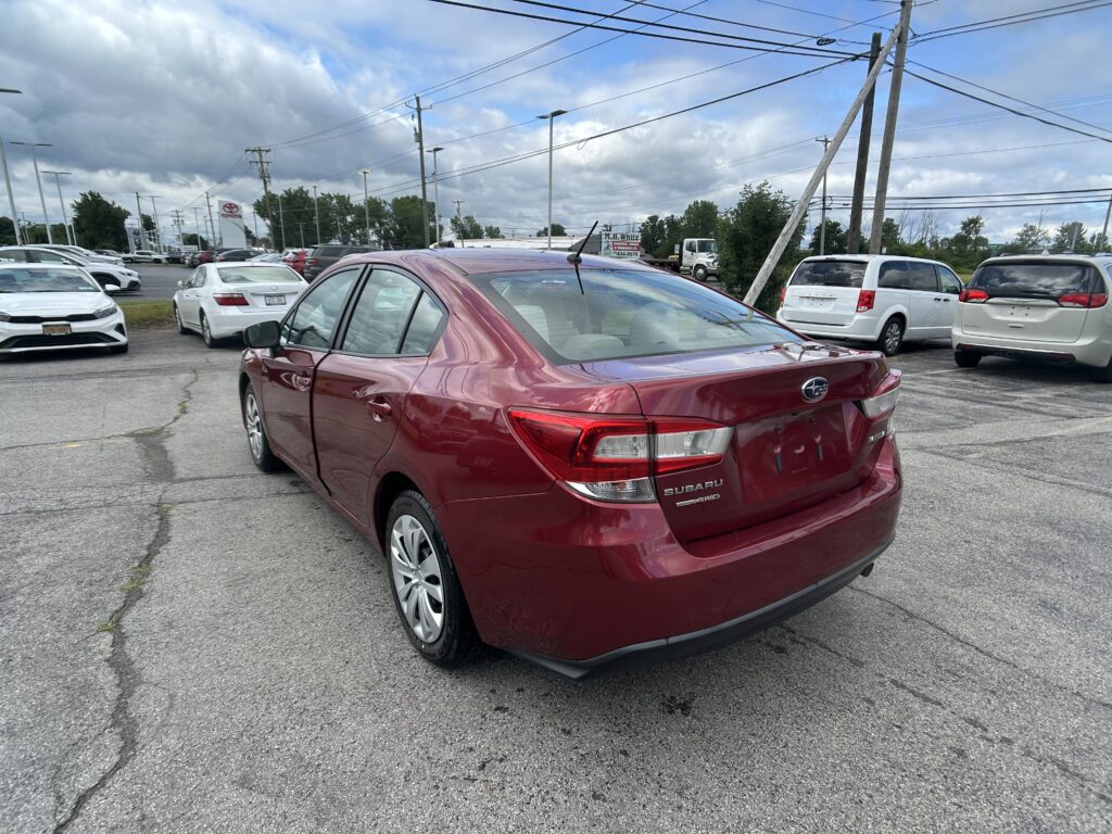 Red Subaru sedan in foreground of a busy car dealership lot with white and silver cars and tall utility poles against a cloudy sky.