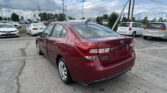 Red Subaru sedan in foreground of a busy car dealership lot with white and silver cars and tall utility poles against a cloudy sky.