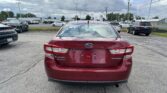 Rear view of a red Subaru Impreza in a car dealership lot, surrounded by parked vehicles and tall light poles under a cloudy sky