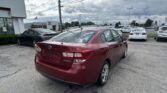 Rear three-quarter view of a maroon Subaru Impreza in a car dealership lot, with a Toyota dealership building and flag in the background under a cloudy sky.