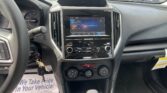 Driver's-side view of a car interior: center touchscreen, climate controls, gear shifter, and keys in the ignition with a floor mat reading 'We Take Pride in Your Vehicle'.","Dashboard interior with touch screen showing FM radio, steering wheel left, and a paper floor mat.","Inside a car: infotainment screen, dashboard vents, gear selector, and a key hanging near the steering column.","Subaru cabin view focusing on center console and instrument cluster, with a printed floor mat in the foreground.","Front-seat view of a vehicle's dashboard and console, including the radio, AC controls, and a promotional paper floor mat."