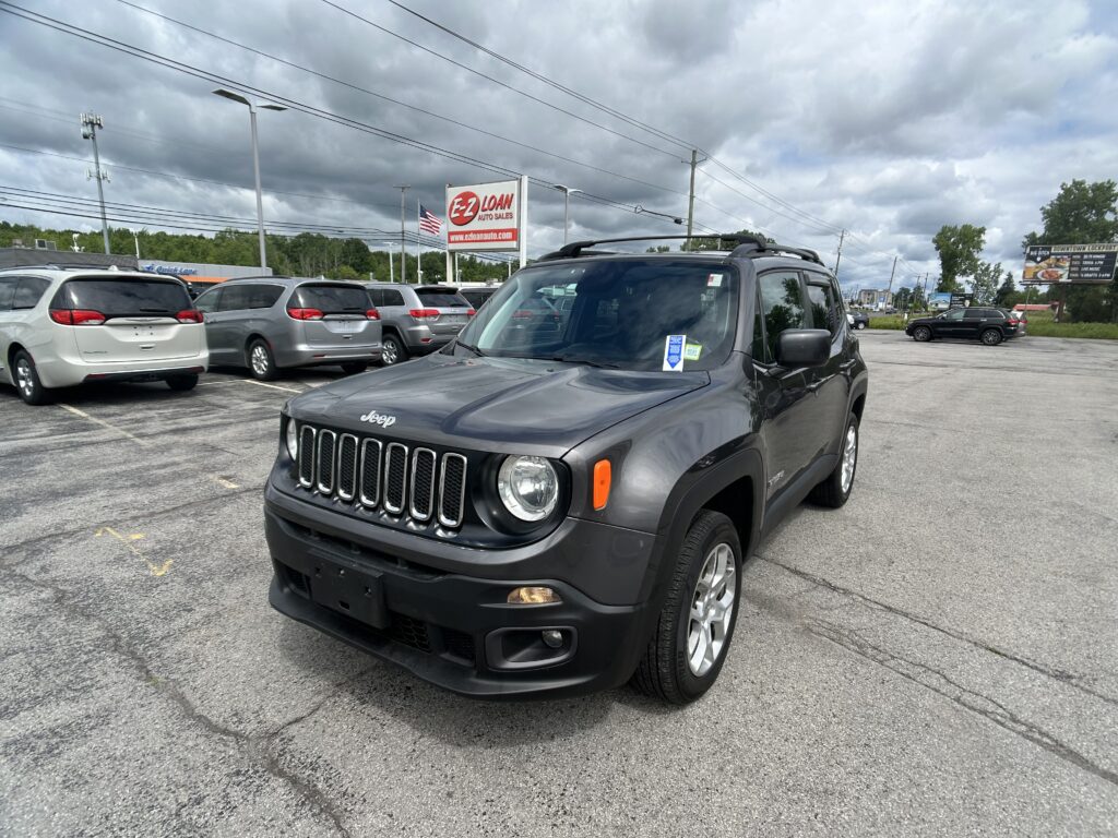 Close-up of a dark gray Jeep SUV in a dealership lot with white minivans in the background and an EZ LOAN Auto Sales sign under a cloudy sky.