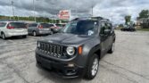 Close-up of a dark gray Jeep SUV in a dealership lot with white minivans in the background and an EZ LOAN Auto Sales sign under a cloudy sky.