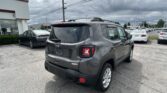 Rear view of a gray Jeep SUV parked in a car dealership lot, with other vehicles and a white building on the left under a cloudy sky.