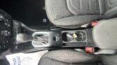 Center console in a car showing gear shifter, climate controls, cup holder, and passenger seat with red seatbelt buckle nearby.