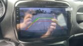In-car touchscreen showing a live rear-view camera view with colored parking guide lines (green, yellow, red) over a parking lot.