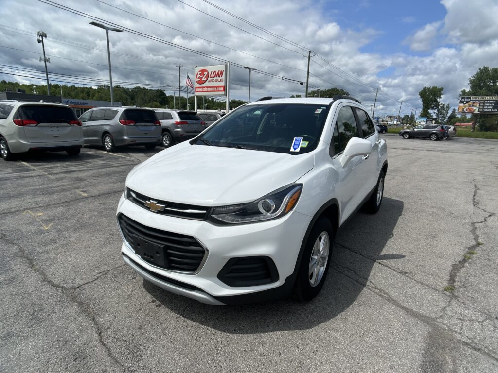 Front-right view of a white Chevrolet SUV in a crowded car lot with multiple minivans nearby and an EZ Loan sign in the background (overcast sky).