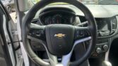 Driver's view of a car interior with a black steering wheel and Chevy emblem, dashboard gauges visible, outside a parking lot full of vehicles. Having the door open on left side.