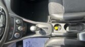 Interior of a car showing the center console with gear shifter, cup holders, and black fabric seats with brown stitching, sunlight casting shadows.