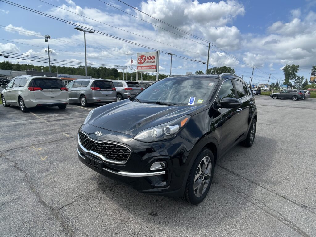 Black Kia SUV in the foreground at a car dealership lot with other minivans nearby under a blue, partly cloudy sky.