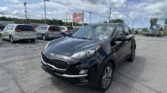Black Kia SUV in the foreground at a car dealership lot with other minivans nearby under a blue, partly cloudy sky.