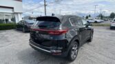 Black Kia SUV shown from the rear three-quarter view in a dealership lot, with other cars and a white building to the left under a cloudy sky.