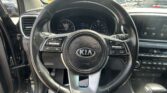 Driver's-eye view of a Kia interior: steering wheel centered, instrument cluster visible, and a large touchscreen showing AV Off, with a parking lot seen through the windshield.