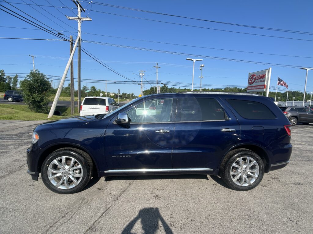 A dark blue SUV parked in a dealership lot with tall power lines and utility poles overhead, and a roadside sign in the background.
