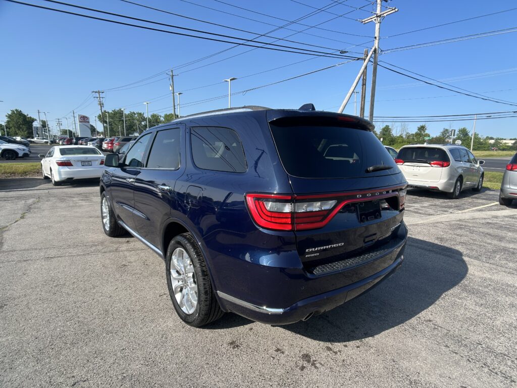Blue Dodge Durango viewed from the rear in a car dealership lot with other vehicles and power lines overhead, clear blue sky behind.