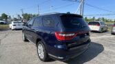 Blue Dodge Durango viewed from the rear in a car dealership lot with other vehicles and power lines overhead, clear blue sky behind.