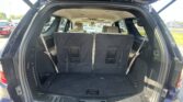 Open SUV tailgate revealing two rows of upright third-row seats and a black carpeted cargo area, with a parking lot visible outside.