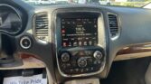 Driver’s-seat view of the dashboard with a touchscreen display showing radio and climate controls; outside is a parking lot full of cars.