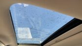 View of blue sky through a car's panoramic sunroof with numerous raindrops on the glass and beige interior framing the opening.
