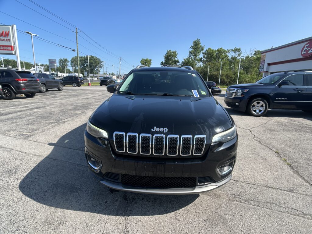 Front view of a black Jeep SUV parked in a busy car dealership lot with other vehicles and signs in the background under a clear blue sky.