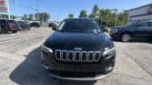 Front view of a black Jeep SUV parked in a busy car dealership lot with other vehicles and signs in the background under a clear blue sky.