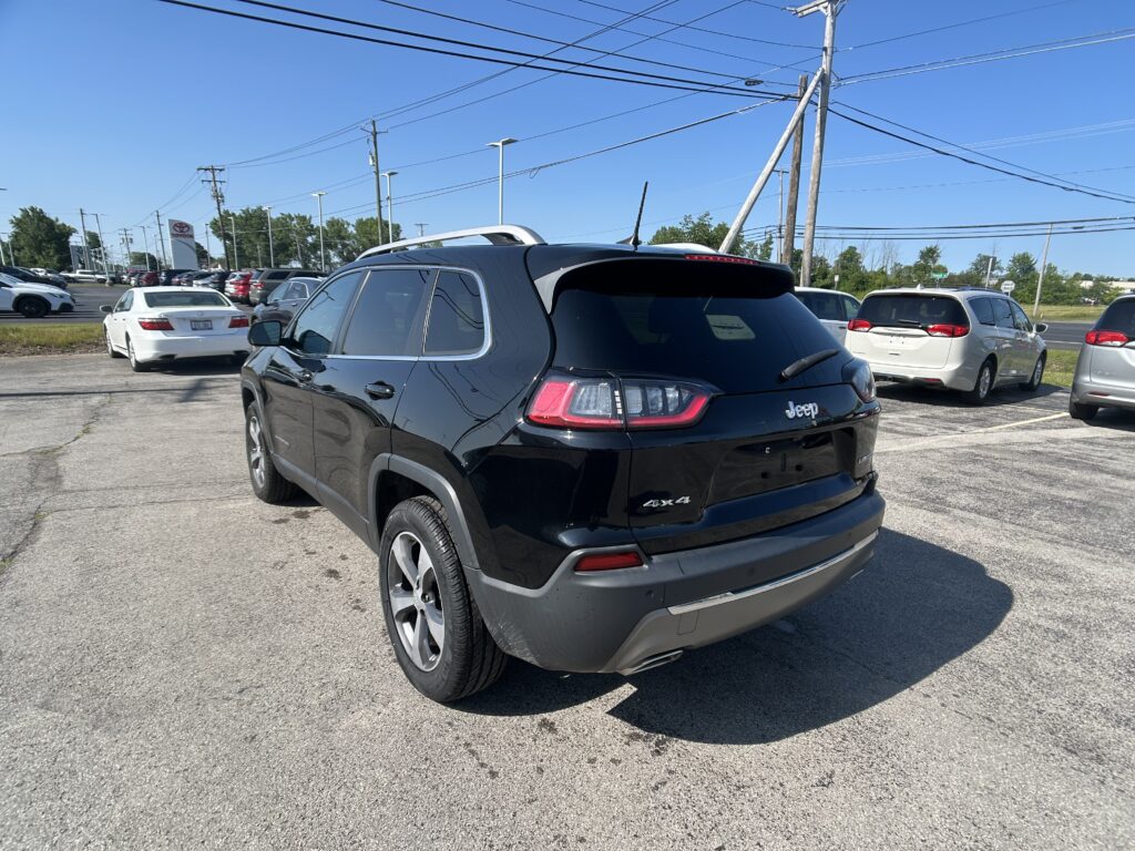 Black Jeep SUV in a car lot, rear three-quarter view, with other vehicles and overhead power lines on a sunny day.