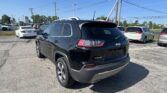 Black Jeep SUV in a car lot, rear three-quarter view, with other vehicles and overhead power lines on a sunny day.