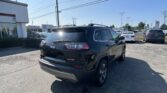 Rear view of a black Jeep SUV in a car dealership lot, with a Toyota dealership and other cars in the background under a clear blue sky