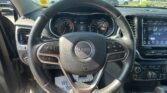 Driver's view of a Jeep steering wheel with the instrument cluster and center touchscreen visible, parked in a dealership lot outside the windshield.”,