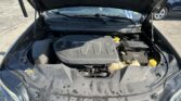 Car engine bay with the hood raised; visible V6 engine cover, battery on the right, and yellow caps in the foreground in a parking lot.