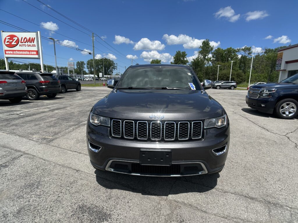 Front view of a dark gray Jeep SUV in a dealership lot, with a bright blue sky and 'E-Z Loan Auto Sales' sign in the background