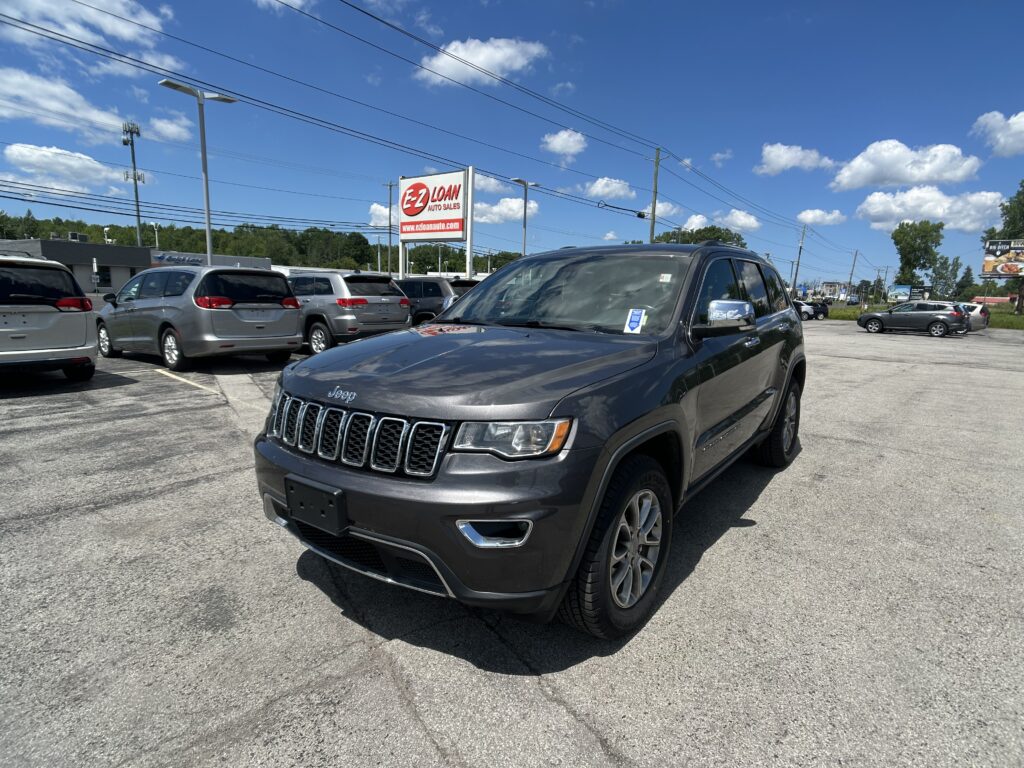 Gray Jeep Grand Cherokee in the foreground at a car dealership, with an EZ Loan Auto Sales sign and blue sky.