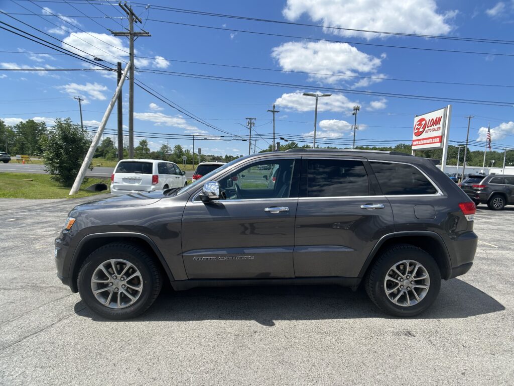 Grey Jeep Grand Cherokee in a dealership lot with power poles and an EZ LOAN Auto Sales sign against a bright blue sky with scattered clouds.