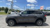 Grey Jeep Grand Cherokee in a dealership lot with power poles and an EZ LOAN Auto Sales sign against a bright blue sky with scattered clouds.