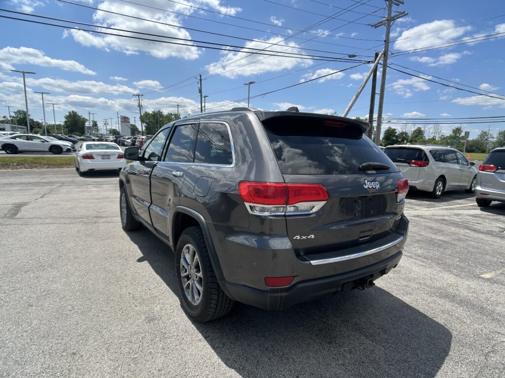 Gray Jeep SUV in foreground at a busy car dealership lot under a blue sky with power lines overhead.