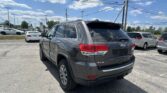 Gray Jeep SUV in foreground at a busy car dealership lot under a blue sky with power lines overhead.