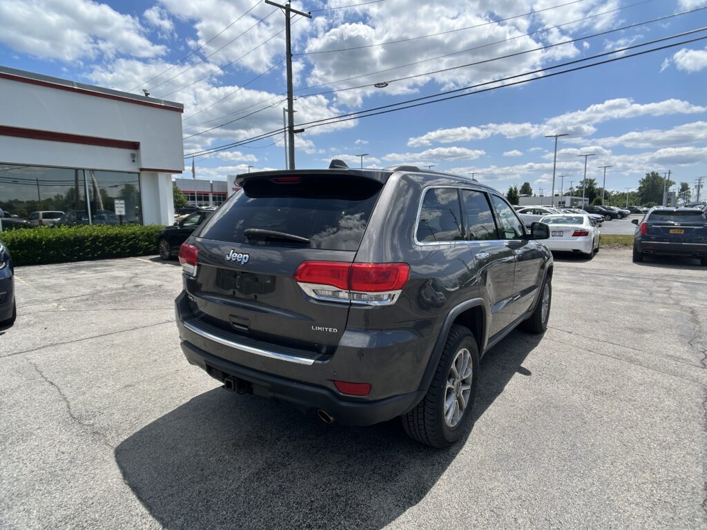 Rear view of a gray Jeep SUV (Limited) in a dealership lot with other cars, glass-front building, and blue sky with clouds above.