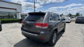 Rear view of a gray Jeep SUV (Limited) in a dealership lot with other cars, glass-front building, and blue sky with clouds above.