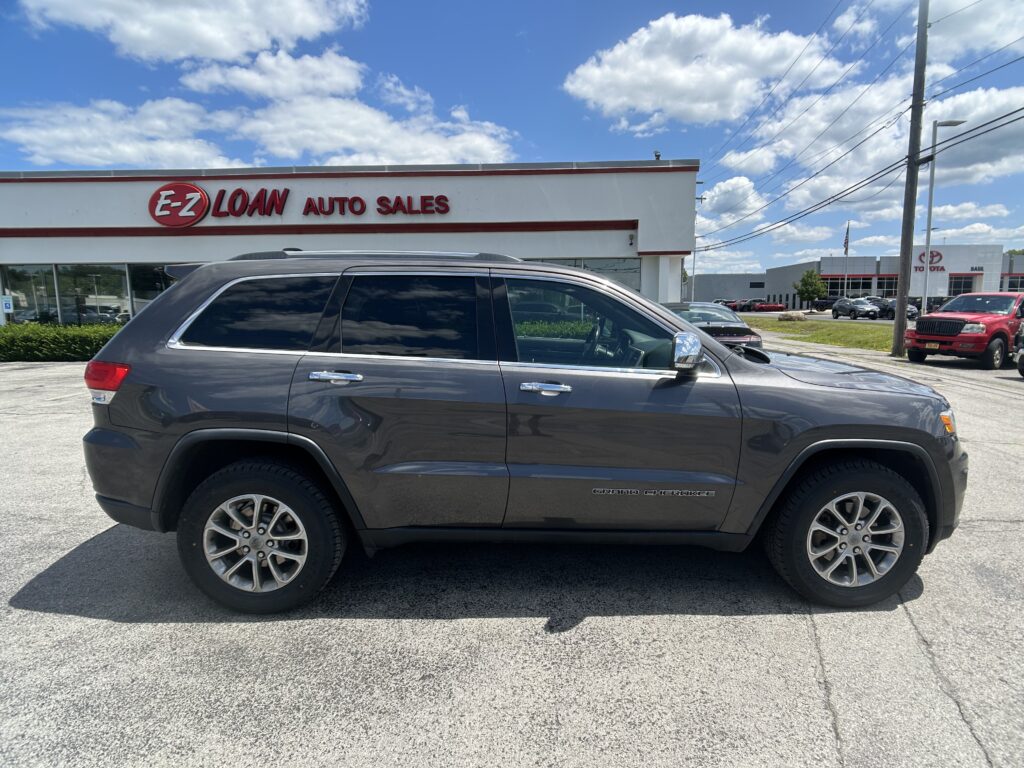 Gray SUV (Jeep Grand Cherokee) parked in front of a white auto dealership building with 'E-Z LOAN Auto Sales' sign on a sunny day