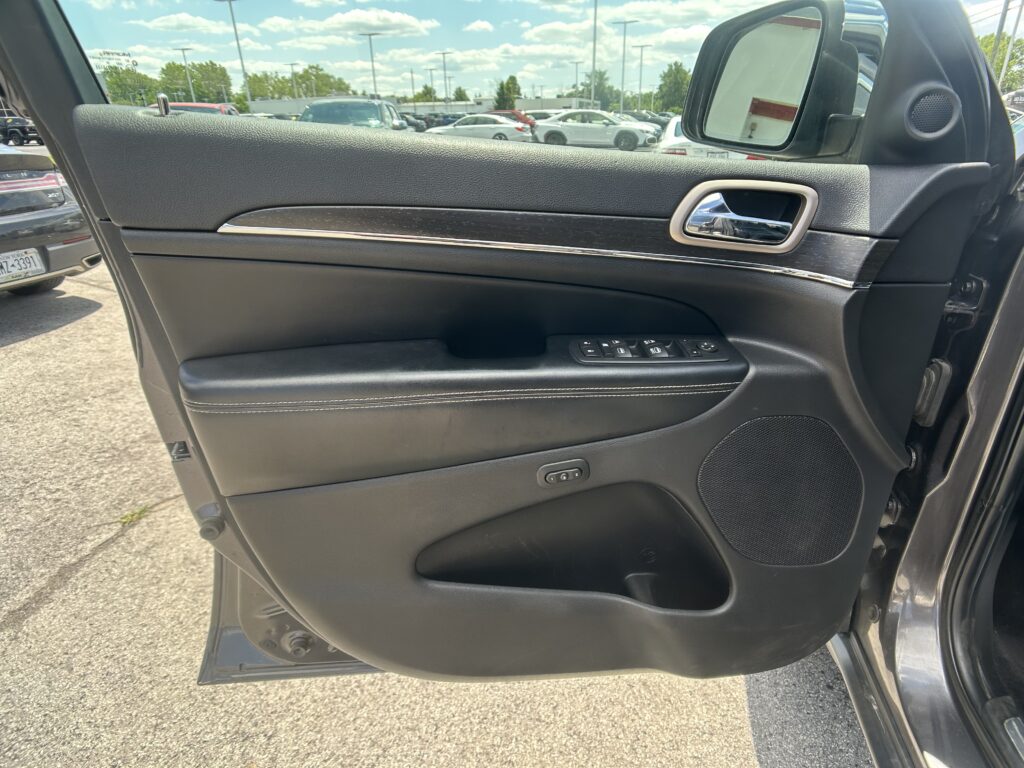 Inside a car door showing the armrest with window controls, chrome door handle, and a speaker, with a parking lot visible outside the window.