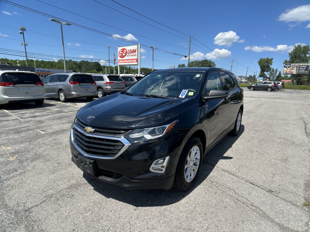 Black Chevrolet SUV in the foreground at a car dealership lot with other minivans and a blue sky above