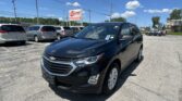 Black Chevrolet SUV in the foreground at a car dealership lot with other minivans and a blue sky above