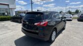 Rear three-quarter view of a black SUV in a car dealership lot, with a white building on the left and other cars parked in the background under a bright blue sky.