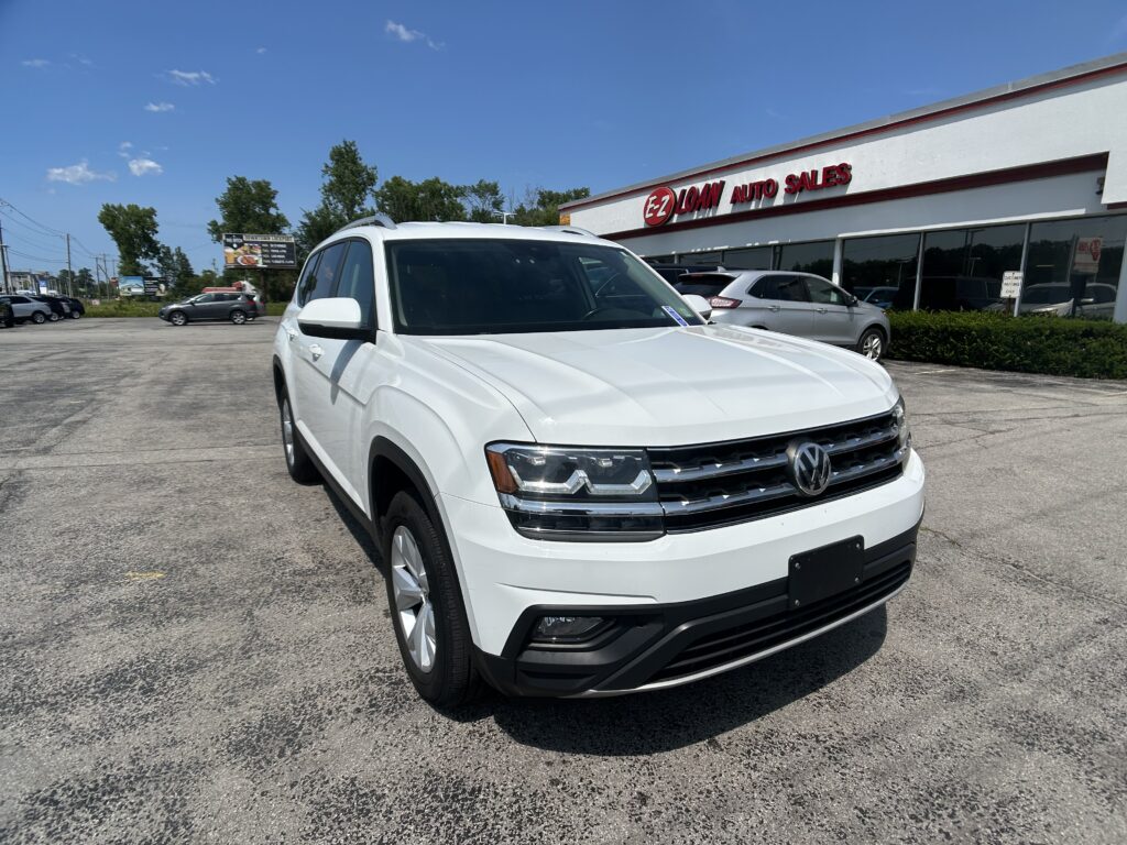 Close-up of a white SUV parked in front of an auto sales lot with a red and white dealership sign behind it on a sunny day.