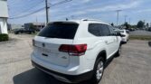 Rear three-quarter view of a white Volkswagen Atlas SUV with roof rails in a dealership parking lot; other vehicles and a service lot visible in the background under a clear blue sky.