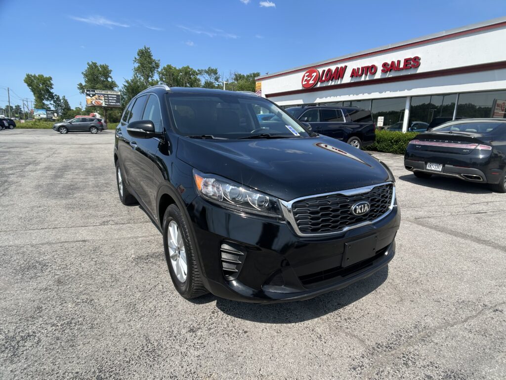 Black Kia SUV in the foreground at a sunny used-car lot, EZ Loan Auto Sales sign visible on the building in the background.