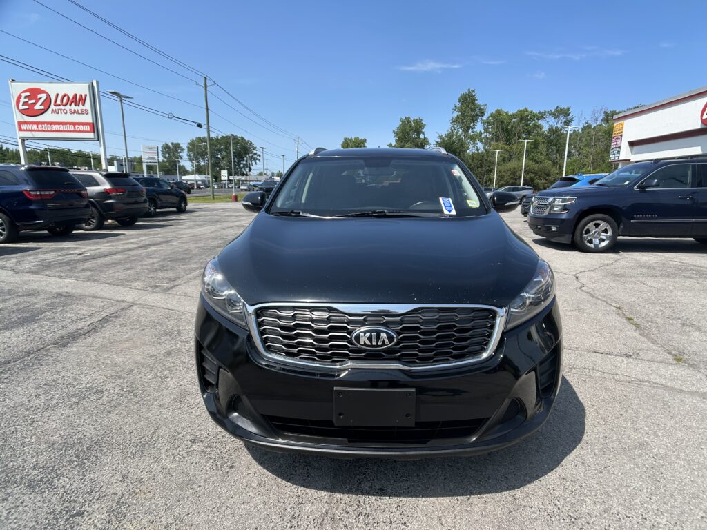 Front view of a black Kia SUV in a dealership lot, with other cars and an EZ Loan Auto Sales sign in the background under a blue sky.