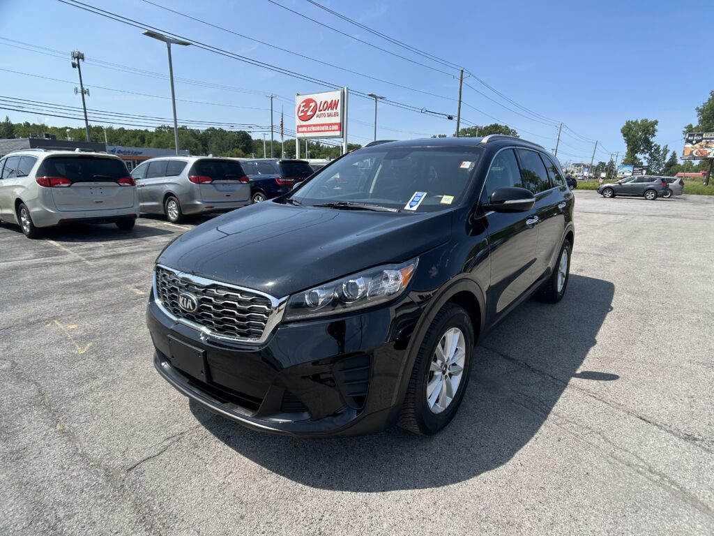 Black Kia SUV in the foreground at a car dealership lot with other minivans and a blue sky in the background, sign reading 'E-Z LOAN Auto Sales'