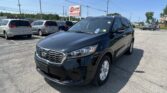 Black Kia SUV in the foreground at a car dealership lot with other minivans and a blue sky in the background, sign reading 'E-Z LOAN Auto Sales'